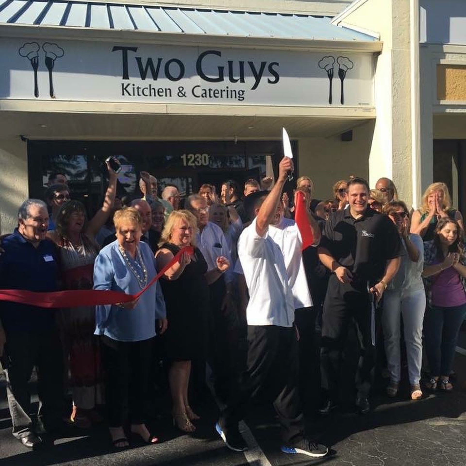 A crowd celebrating a ribbon cutting outside Two Guys Kitchen and Catering