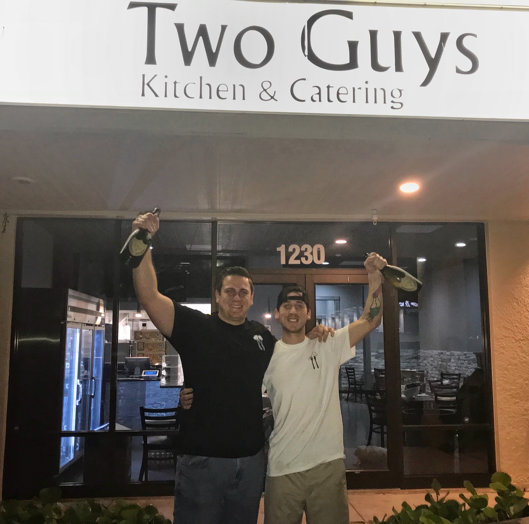 Two men raising bottles in celebration outside the Two Guys Kitchen and Catering storefront